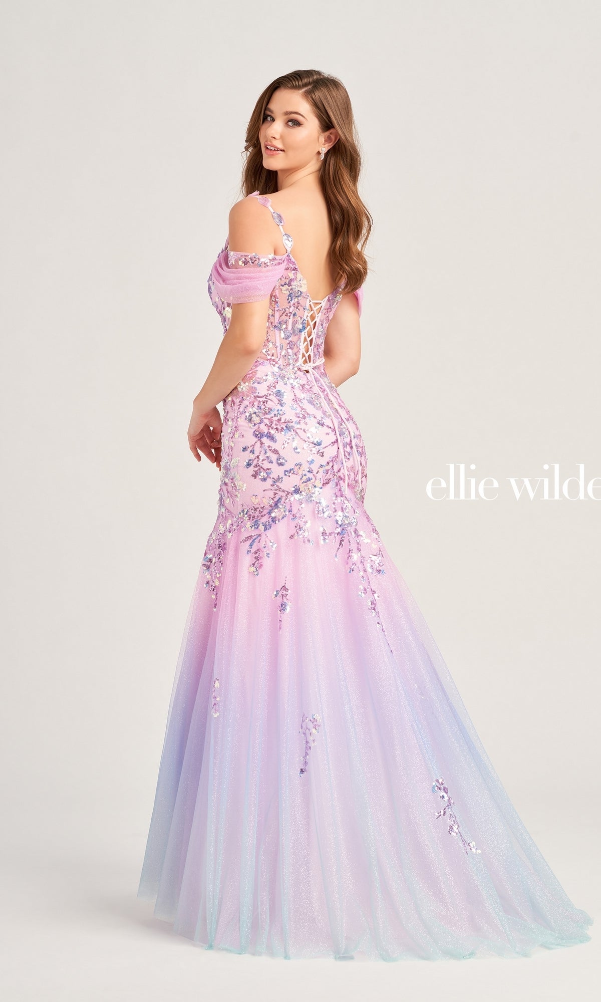 A woman poses in the Cold-Shoulder Ellie Wilde Long Prom Dress EW35056, featuring floral embellishments, off-the-shoulder sleeves, and a lace-up back, in front of a plain light background.